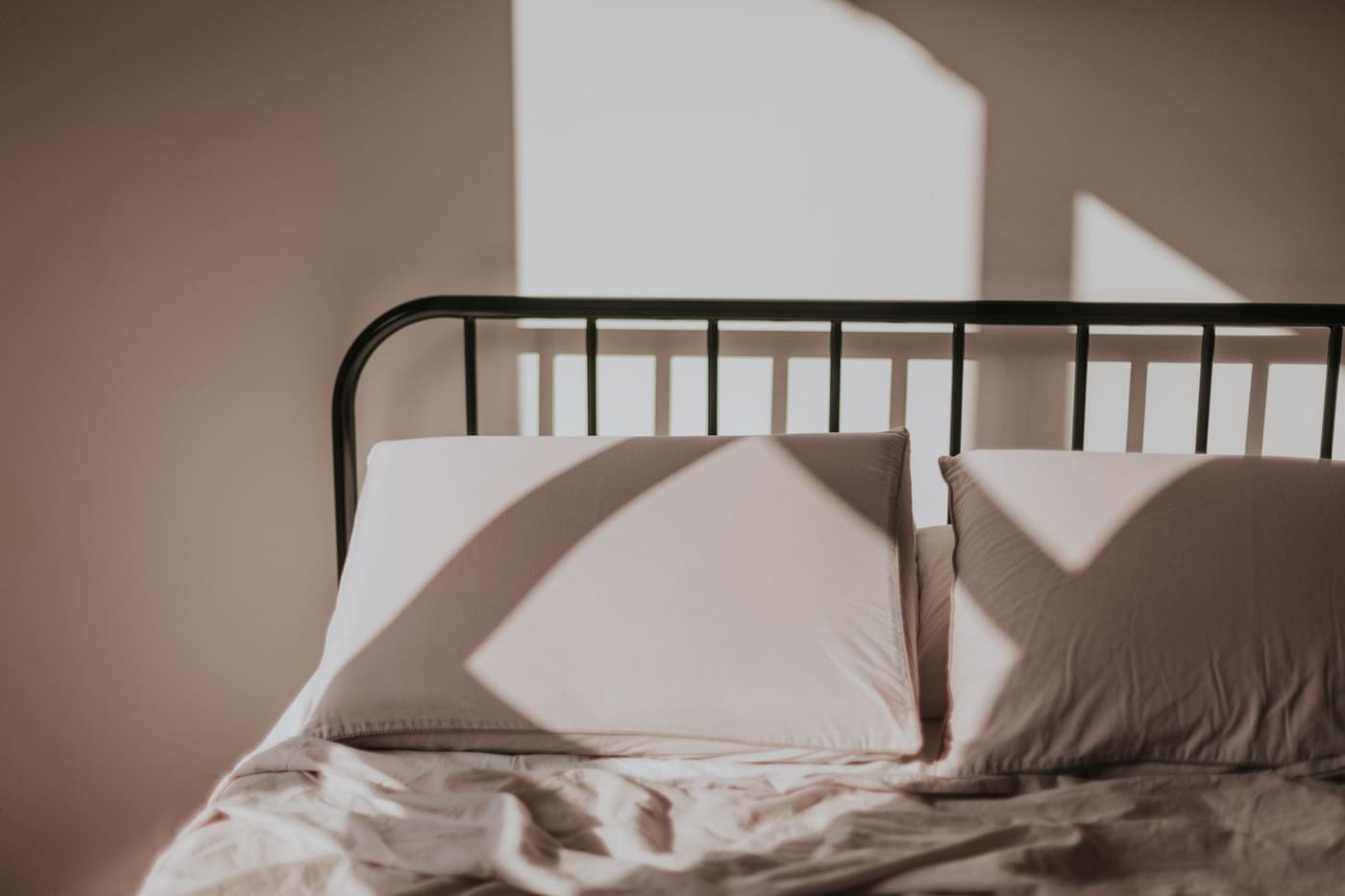 A bed with white pillows against a dark metal headboard and sunlight casting window shadows on the wall