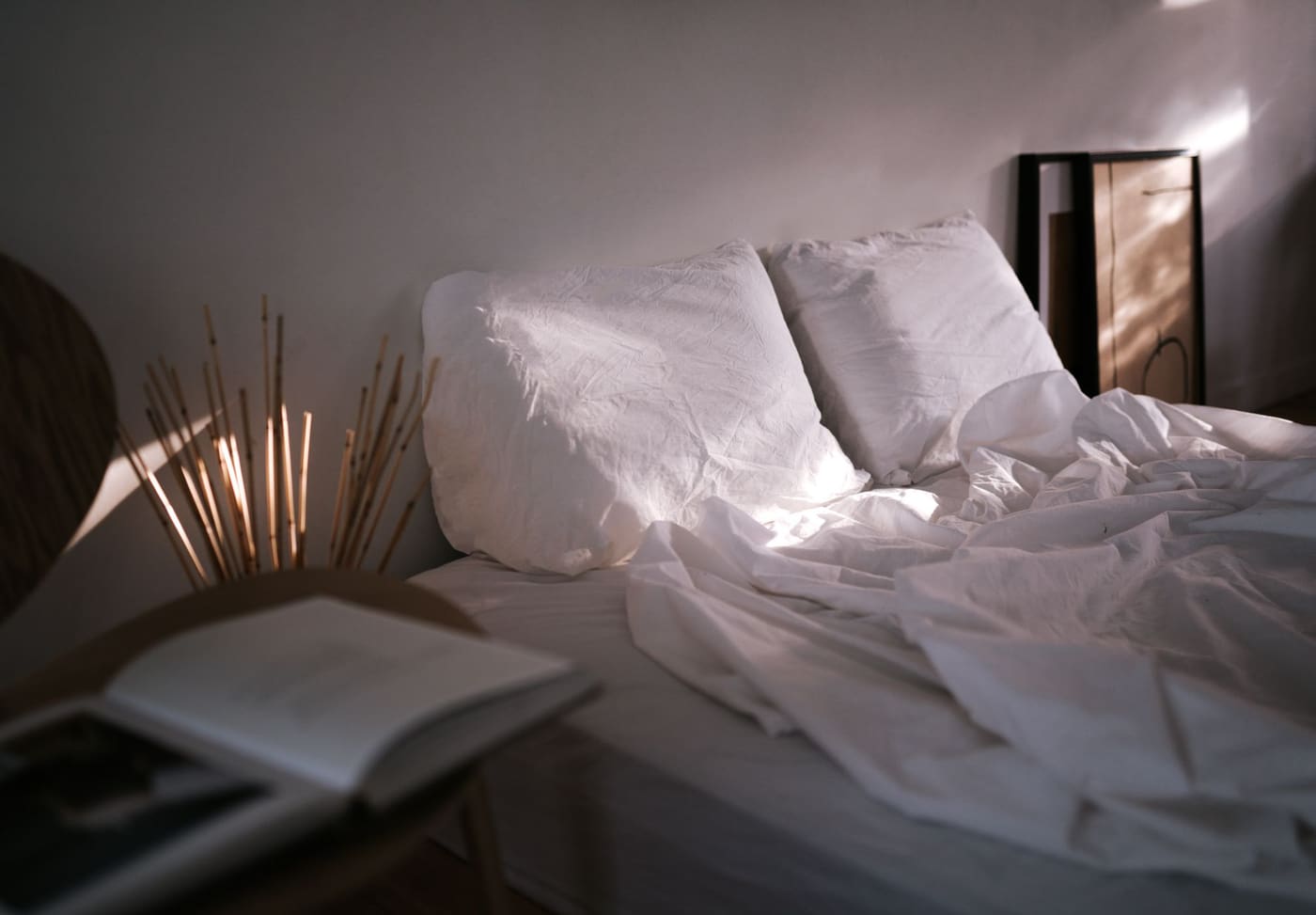 An unmade bed with white linen and pillows, a book and dried reeds on the nightstand