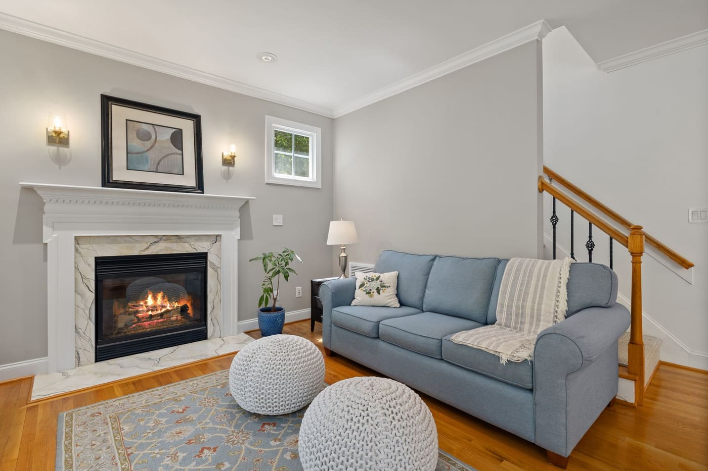 A living room with a blue sofa, a lit fireplace, a patterned rug and two knitted poufs