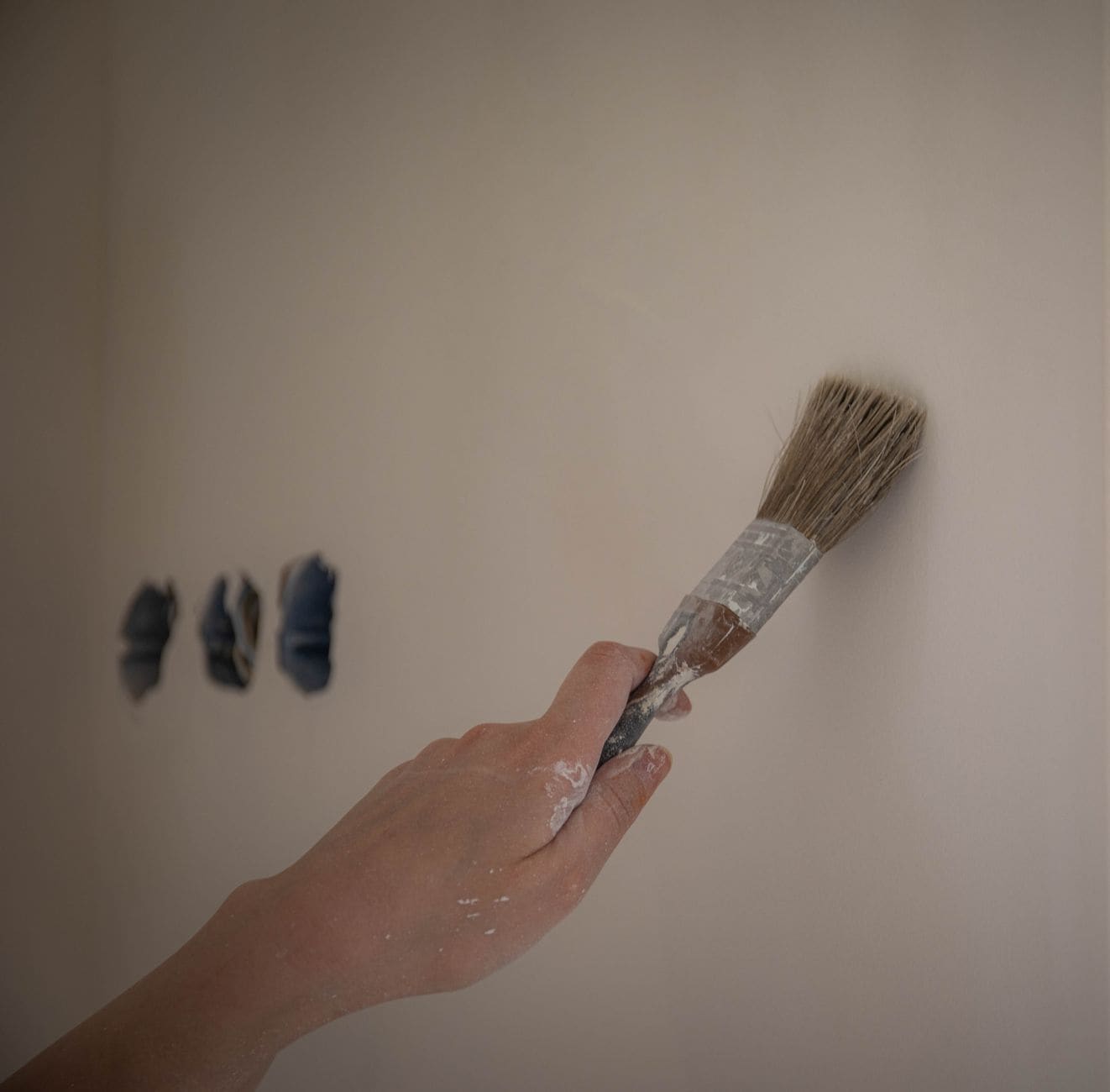 A hand using a brush to test three small paint patches on a pale wall