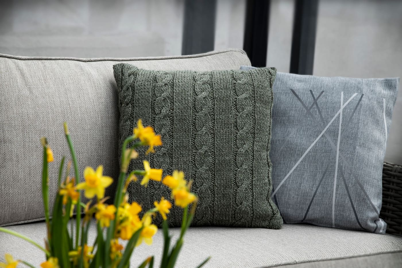 A gray sofa with knitted and linen pillows and a bunch of yellow daffodils in front