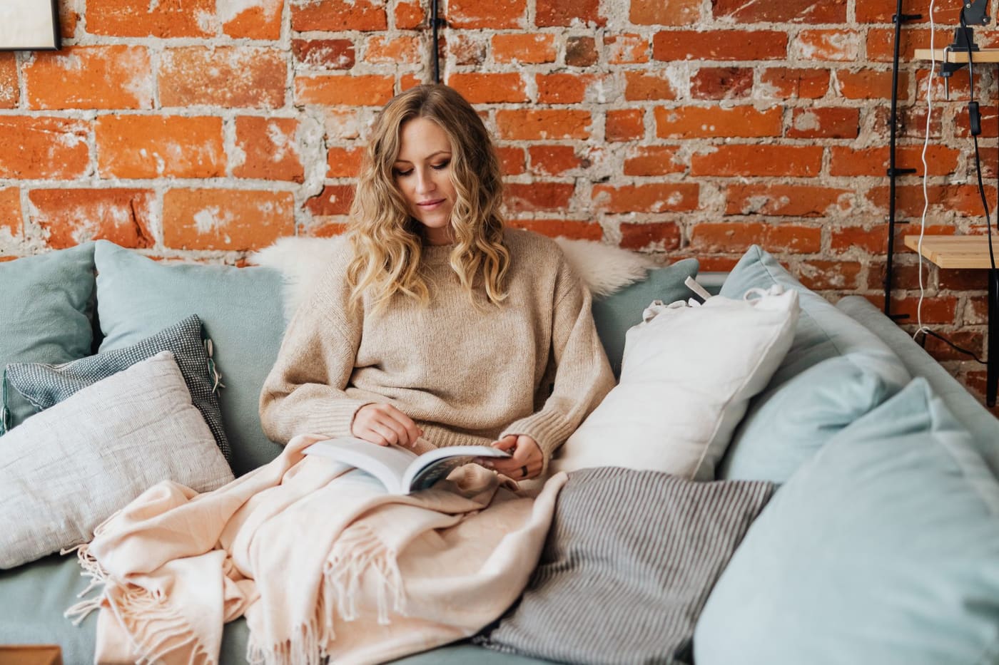A person reading on a sofa under a light knit blanket in front of a brick wall