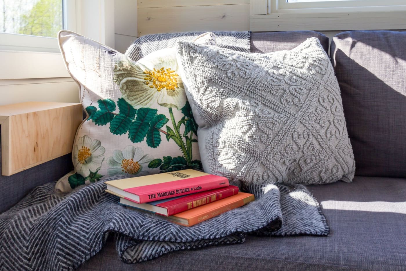 A sunlit sofa corner with decorative pillows, a stack of books and a blanket