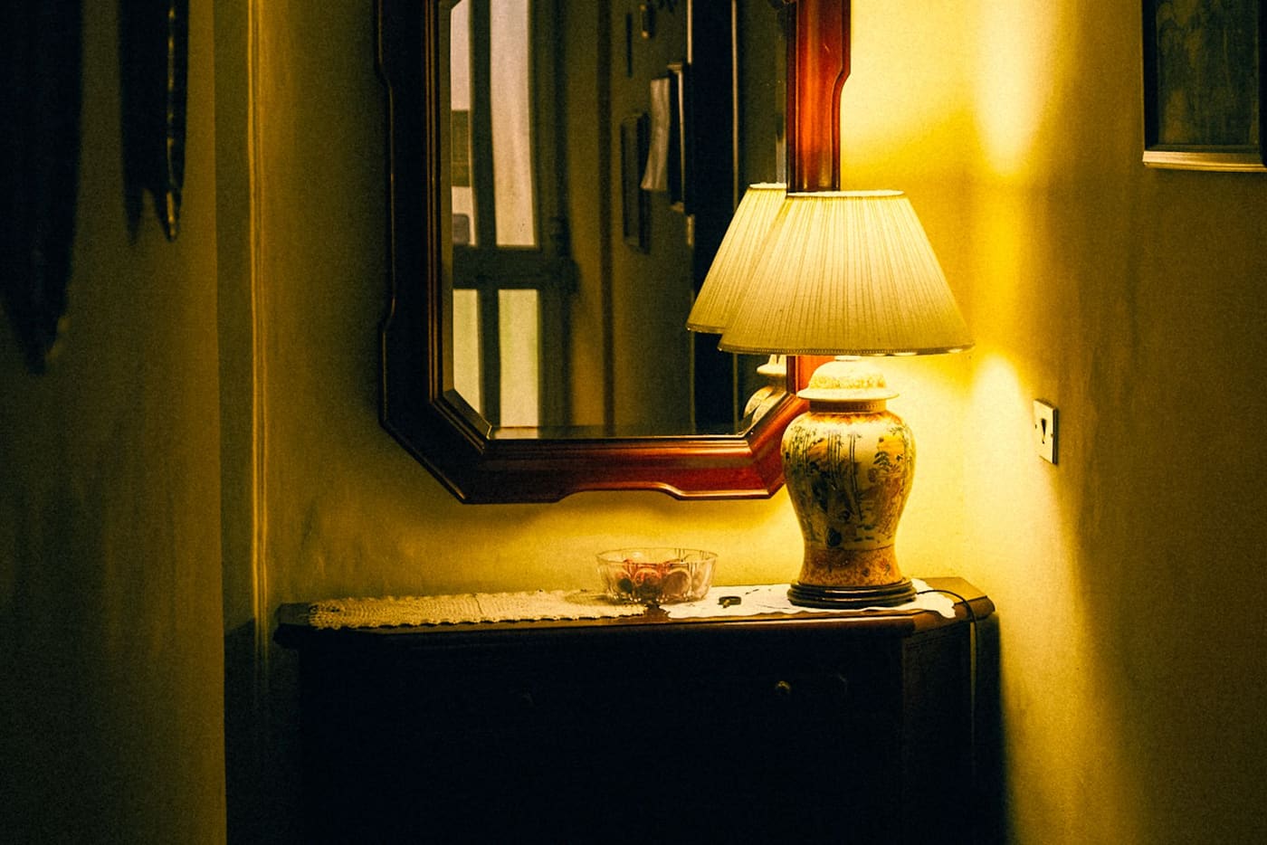 A table lamp with a pale fabric shade glowing on a console table beside a framed mirror