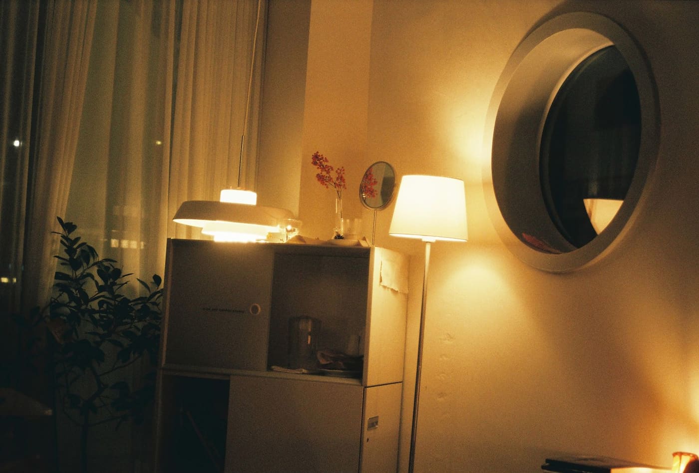 An evening room lit by a floor lamp and several warm lights, with a round mirror and plants